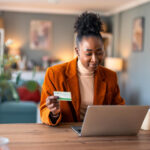 Woman shopping online on her laptop while holding a credit card at her desk.